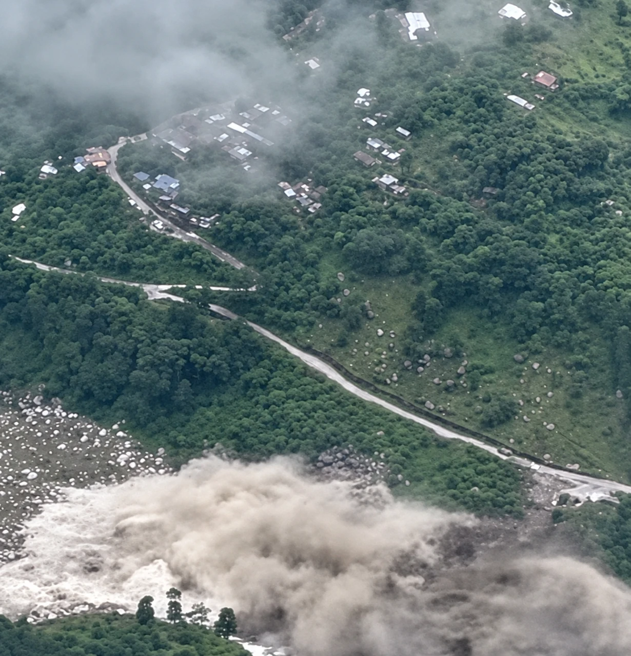 Aerial view of a hillside village partly covered by cloud, with a road leading down to a river carrying floodwater and debris