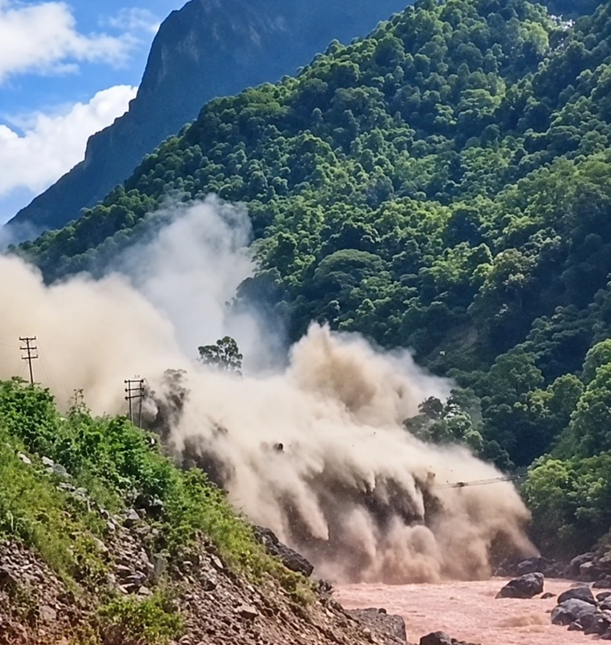 A large cloud of dust and debris rising from a river gorge surrounded by forested mountains